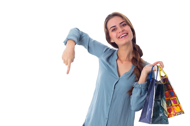 Jovem sorridente de blusa azul segurando sacolas de compras coloridas e apontando para baixo no estúdio