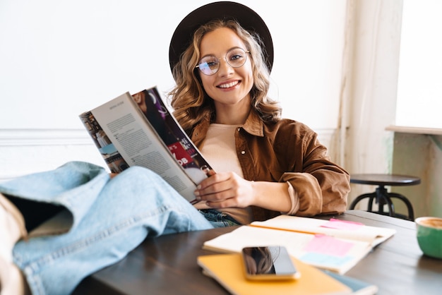 Jovem sorridente de chapéu lendo revista com as pernas na mesa enquanto estudava em casa