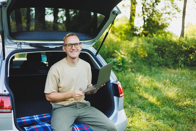 Jovem sorridente de óculos freelancer trabalha em um laptop enquanto está sentado no porta-malas de um carro Trabalho remoto na natureza