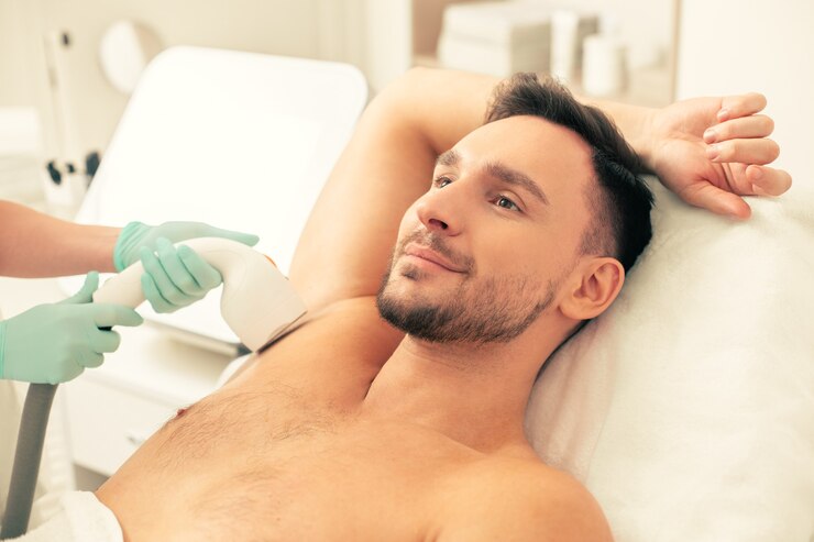 Jovem sorridente deitado no travesseiro com uma mão atrás da cabeça e fazendo uma depilação a laser na axila