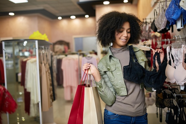 Jovem sorridente e atraente mulher afro-americana escolhendo o tamanho certo do sutiã na boutique da loja de lingerie