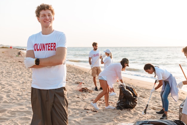 Jovem sorridente e confiante voluntário limpando o lixo da praia com um grupo de voluntários