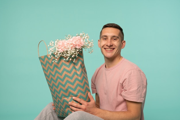 Jovem sorridente e feliz sentado no chão, segurando uma sacola de compras com flores