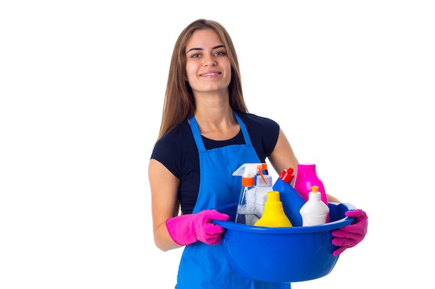 Jovem sorridente em camiseta azul e avental com luvas segurando coisas de limpeza na bacia azul