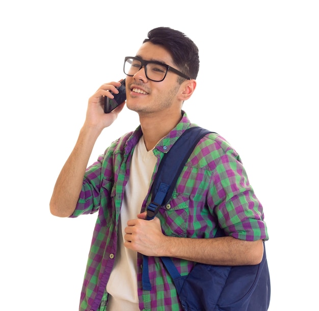 Jovem sorridente em camiseta branca e camisa quadriculada com mochila azul falando em seu smartphone