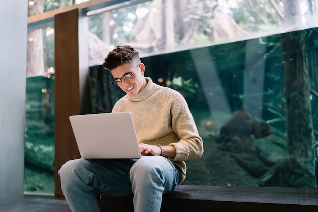 Jovem sorridente em óculos digitando no laptop perto do aquário