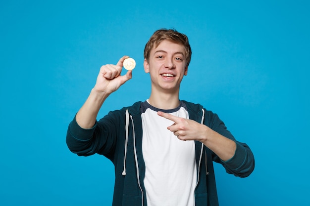 Jovem sorridente em roupas casuais, apontando o dedo indicador na moeda futura de bitcoin isolada na parede azul. Emoções sinceras de pessoas, conceito de estilo de vida.