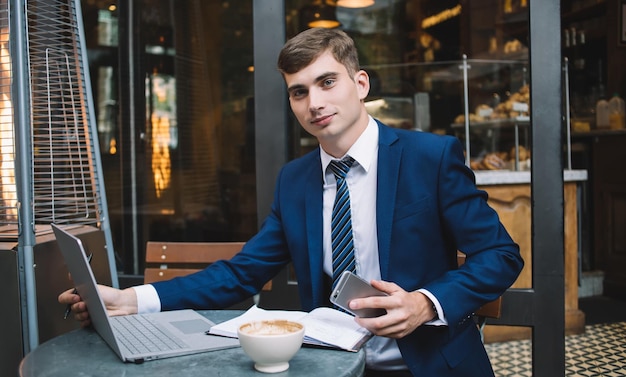 Jovem sorridente em terno de negócios com smartphone sentado na mesa com notebook laptop e xícara de café enquanto olha para a câmera no café