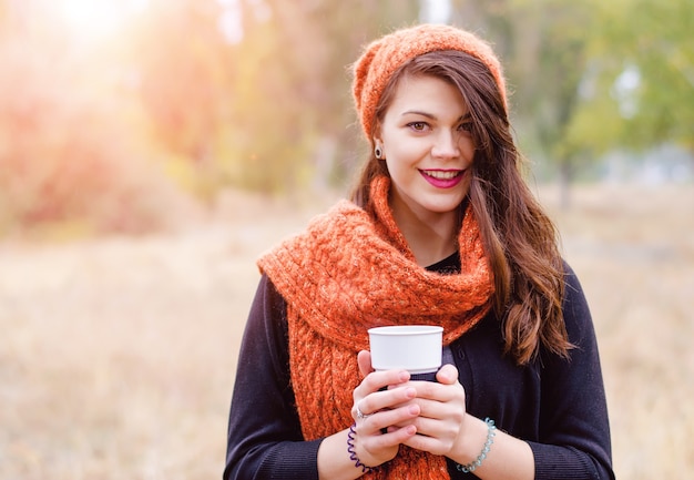 Jovem sorridente em um lenço e um chapéu com uma xícara de café (chá) nas mãos do outono ao ar livre. Os raios do sol iluminam o modelo por trás.