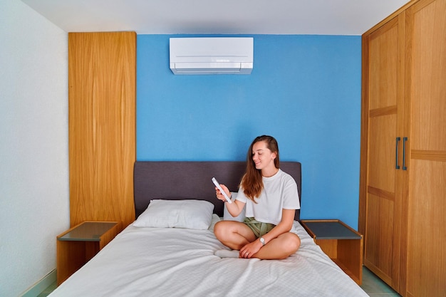 Jovem sorridente feliz e fofa sentada na cama e ajustando a temperatura do ar condicionado usando controle remoto no quarto