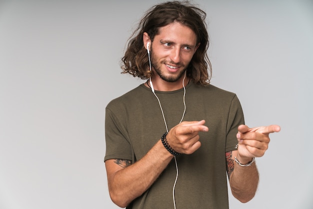 Jovem sorridente gosta de ouvir música com fones de ouvido em pé e apontando os dedos para o espaço da cópia isolado em cinza