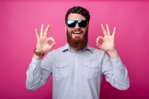 Jovem sorridente homem barbudo com óculos de sol, mostrando o gesto OK