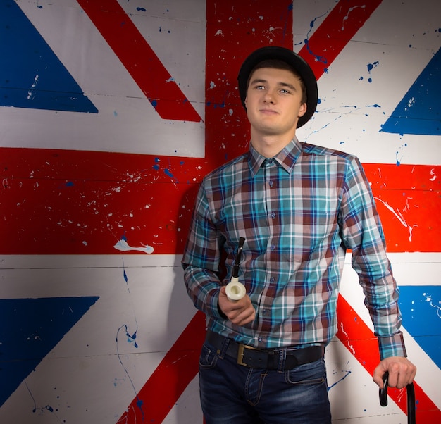 Jovem sorridente homem bonito com cachimbo e bengala em roupa da moda na frente da bandeira da Grã-Bretanha