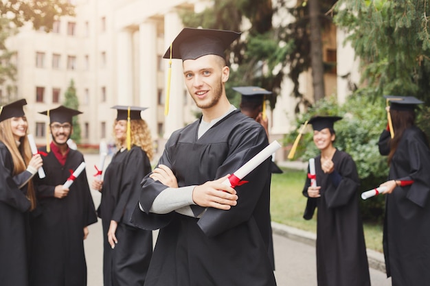 Jovem sorridente masculino em seu dia de formatura na universidade em pé com grupo multiétnico de pessoas. Educação, qualificação e conceito de vestido.