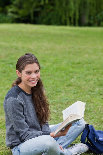 Jovem, sorridente, menina, sentada, de pernas cruzadas, segurando um livro