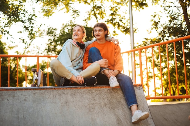 Foto jovem sorridente menino e menina olhando alegremente de lado enquanto passam tempo juntos no skatepark