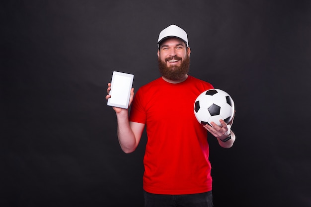 jovem sorridente, mostrando o tablet e segurando uma bola de futebol sobre o preto