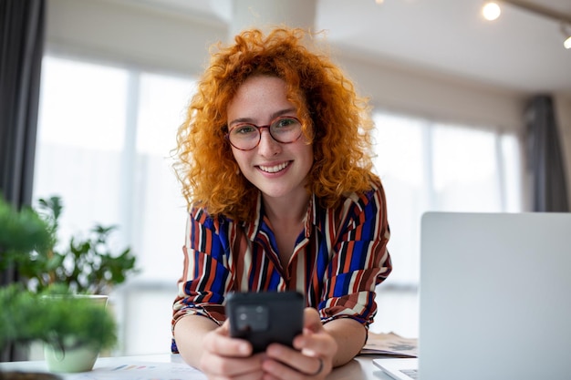 Jovem sorridente mulher de negócios de cabelo vermelho usando smartphone perto de lptop no escritório