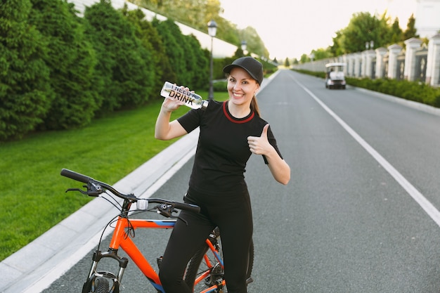 Jovem sorridente mulher forte atlética em uniforme preto, boné segurando apontando na garrafa de água, andando de bicicleta ao ar livre em dia ensolarado de primavera ou verão. fitness, esporte, conceito de estilo de vida saudável.