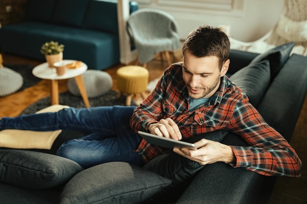 Jovem sorridente relaxando em casa e navegando na Internet no touchpad