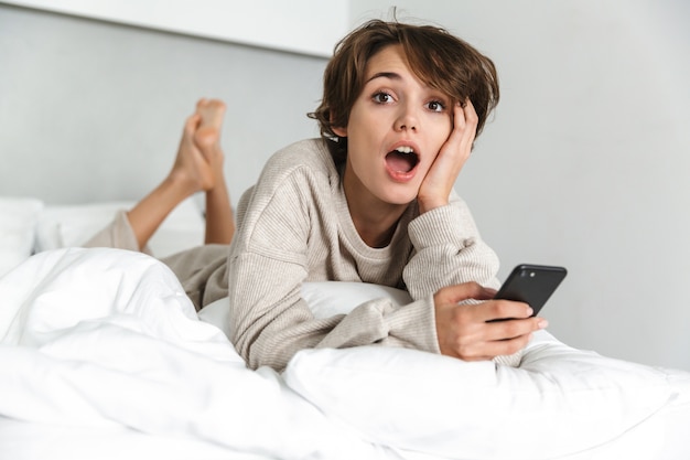 Jovem sorridente relaxando na cama pela manhã, usando um telefone celular