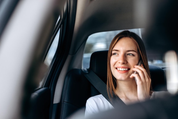 Jovem sorridente segurando o smartphone na mão e falando no banco de trás do carro Senhora muito feliz tendo viagem de negócios