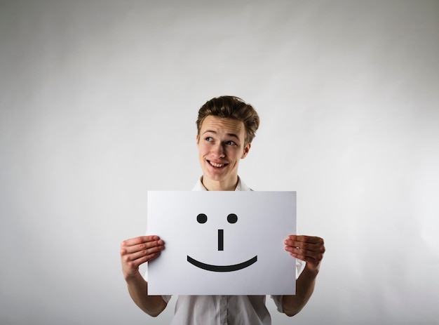 Foto jovem sorridente segurando papel com rosto antropomórfico enquanto está de pé contra um fundo cinzento