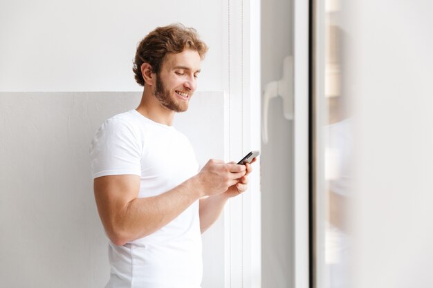 Jovem sorridente segurando um telefone celular, em frente à janela de casa