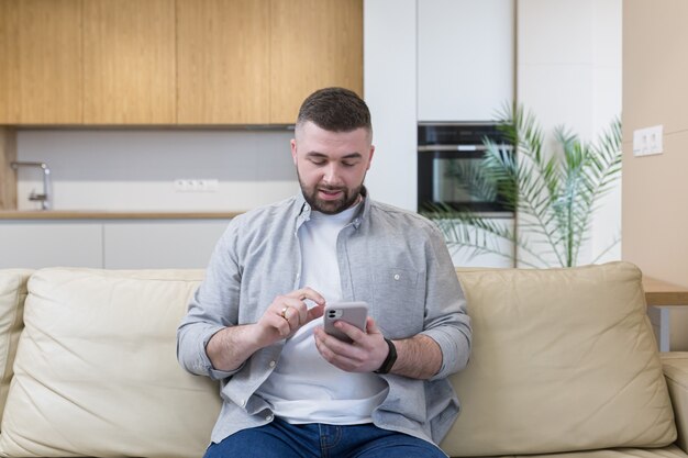 Jovem sorridente segurando um telefone celular enquanto está sentado em um sofá em casa e comemorando