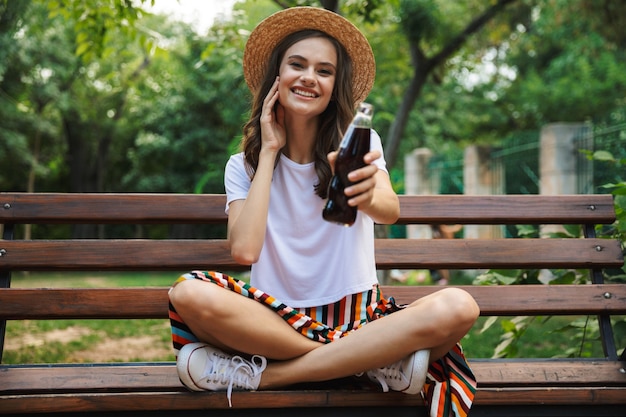 Jovem sorridente segurando uma garrafa com refrigerante enquanto está sentado no parque ao ar livre