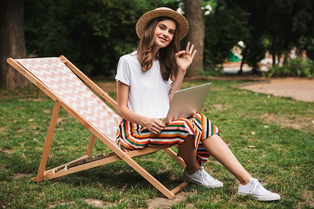 Jovem sorridente sentada com um laptop no parque ao ar livre, mostrando-se bem