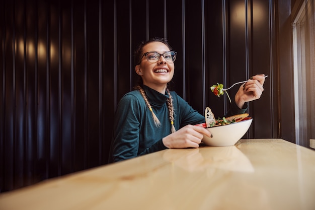 Jovem sorridente sentada em um restaurante ao lado da janela e comendo salada no almoço. Estudante, faculdade