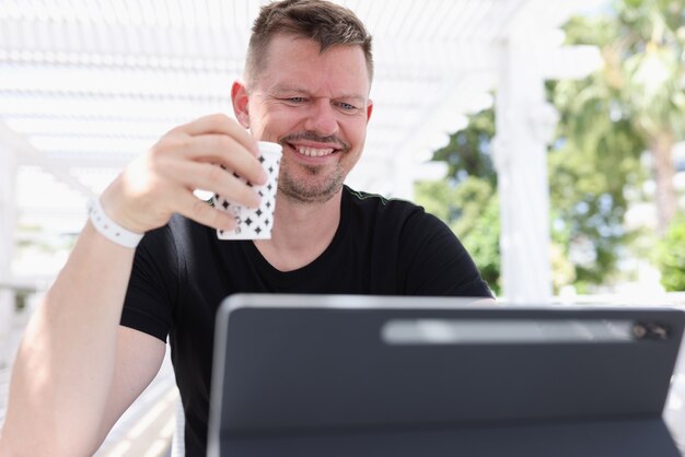 Jovem sorridente sentado com uma xícara de café e trabalhando em um tablet na rua