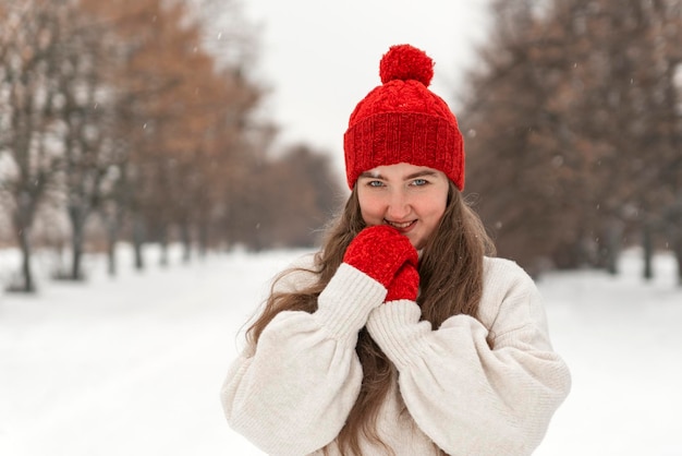 Jovem sorridente tímida com chapéu de malha vermelho e luvas no parque de inverno nevado Retrato de jovem caminhando