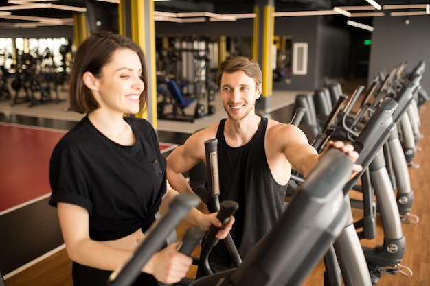 Jovem sorridente treinando no aparelho elíptico com instrutor de fitness em pé perto dela e obs