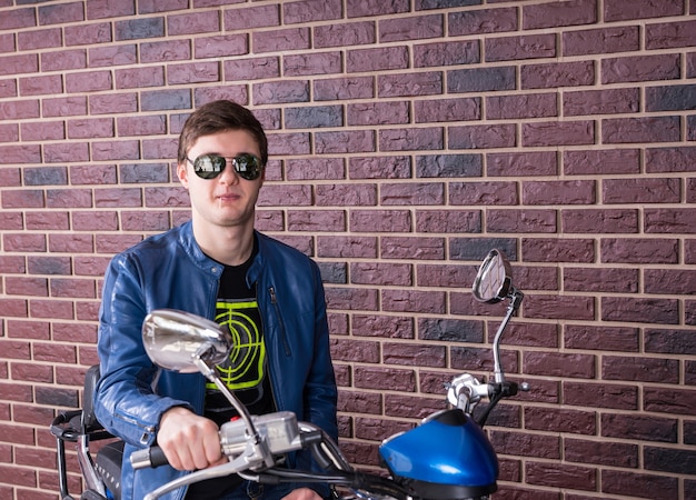 Jovem sorridente, usando óculos escuros da moda, sentado e relaxando em uma moto azul em frente a uma parede de tijolos, sorrindo para a câmera