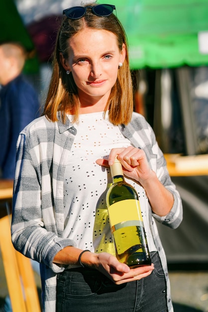 Jovem sorridente usando óculos escuros detém a garrafa de vinho. Estilo casual de mulher. Pessoa do sexo feminino se divertindo. Boa celebração do feriado. Retrato de estilo de vida ao ar livre. Rosto bonito adulto. Evento de rua