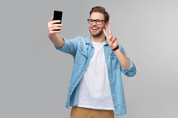 Jovem sorridente, vestindo uma camisa jeans, tirando uma foto de selfie no smartphone ou fazendo uma videochamada em pé sobre a parede cinza do estúdio