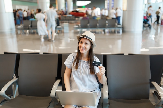 Jovem sorridente viajante turista mulher usando chapéu, trabalhando em um laptop, segurando um cartão de crédito, esperando no saguão do aeroporto internacional