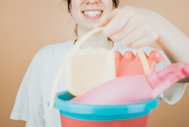 Jovem sorrindo alegremente para a câmera enquanto segura o cubo com brinquedos para brincar na praia ao ar livre no verão Imagem de fundo colorida