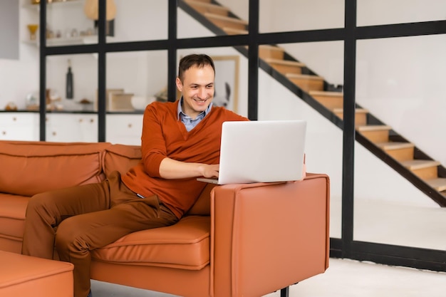 Jovem sorrindo, lê a tela de um computador portátil enquanto relaxa em um sofá confortável em casa.