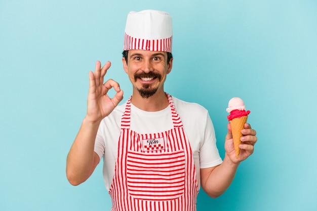 Jovem sorveteira caucasiana segurando um sorvete isolado em um fundo azul alegre e confiante mostrando um gesto ok.
