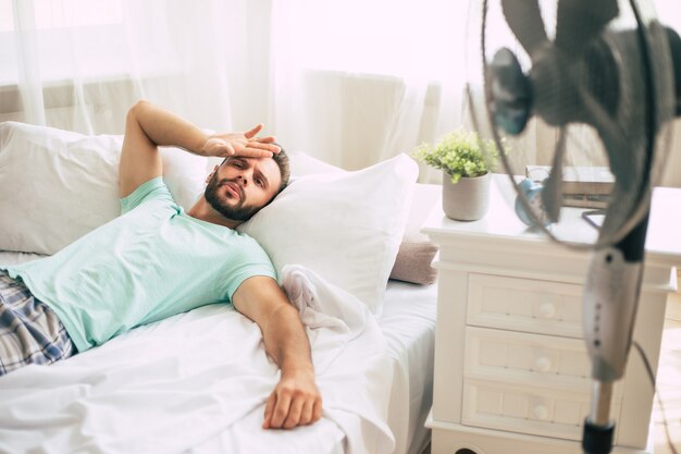 Jovem suado está tentando se refrescar do calor com um ventilador enquanto está deitado na cama em casa.