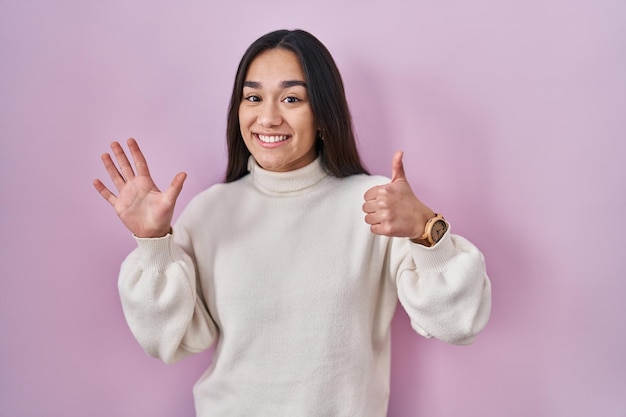 Jovem sul-asiática em pé sobre fundo rosa mostrando e apontando para cima com os dedos número seis enquanto sorria confiante e feliz