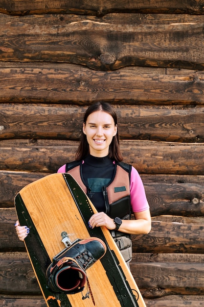 Jovem surfista ativa com jaqueta de segurança, segurando a prancha de surf, enquanto fica de pé contra a parede de uma casa de madeira