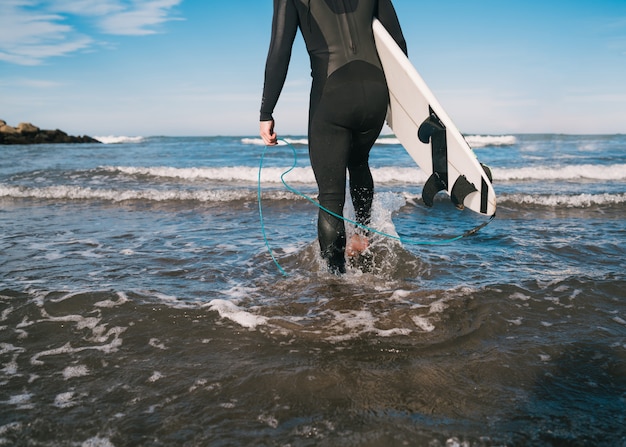 Jovem surfista entrando na água com sua prancha de surf em um traje de surf preto. conceito de esporte e esporte de água.