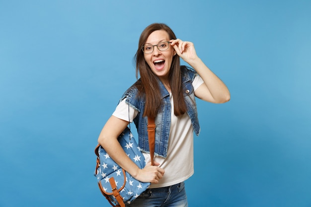 Jovem surpreendeu a estudante feliz com a boca aberta em uma camiseta branca e roupas jeans com mochila segurando óculos isolados sobre fundo azul. Educação no conceito de faculdade de Universidade de ensino médio.