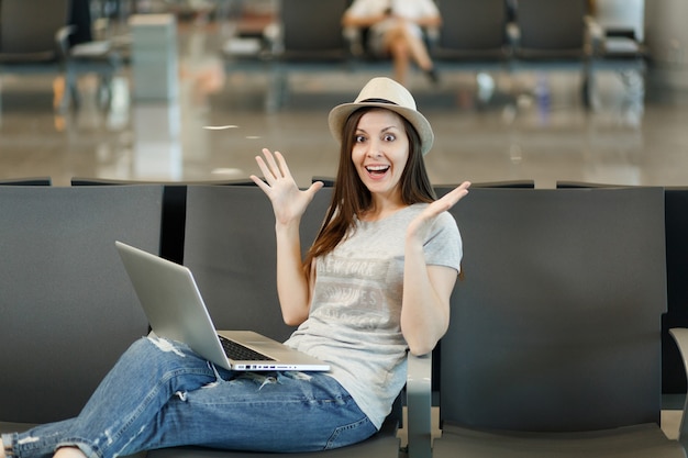 Jovem surpreendeu a mulher turista viajante sentada, trabalhando em um laptop, espalhando as mãos, esperando no saguão do aeroporto internacional