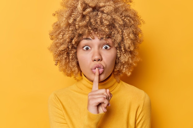 Jovem surpreendida com cabelo espesso encaracolado faz gesto de silêncio compartilha segredo mantém os olhos amplamente abertos usa jumper casual isolado sobre fundo amarelo vívido Conceito de pessoas e sigilo