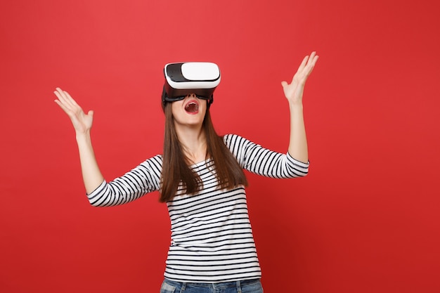 Jovem surpreendida em roupas listradas, óculos de realidade virtual, mantendo a boca aberta, espalhando as mãos isoladas no fundo da parede vermelha. conceito de estilo de vida de emoções sinceras de pessoas. simule o espaço da cópia.